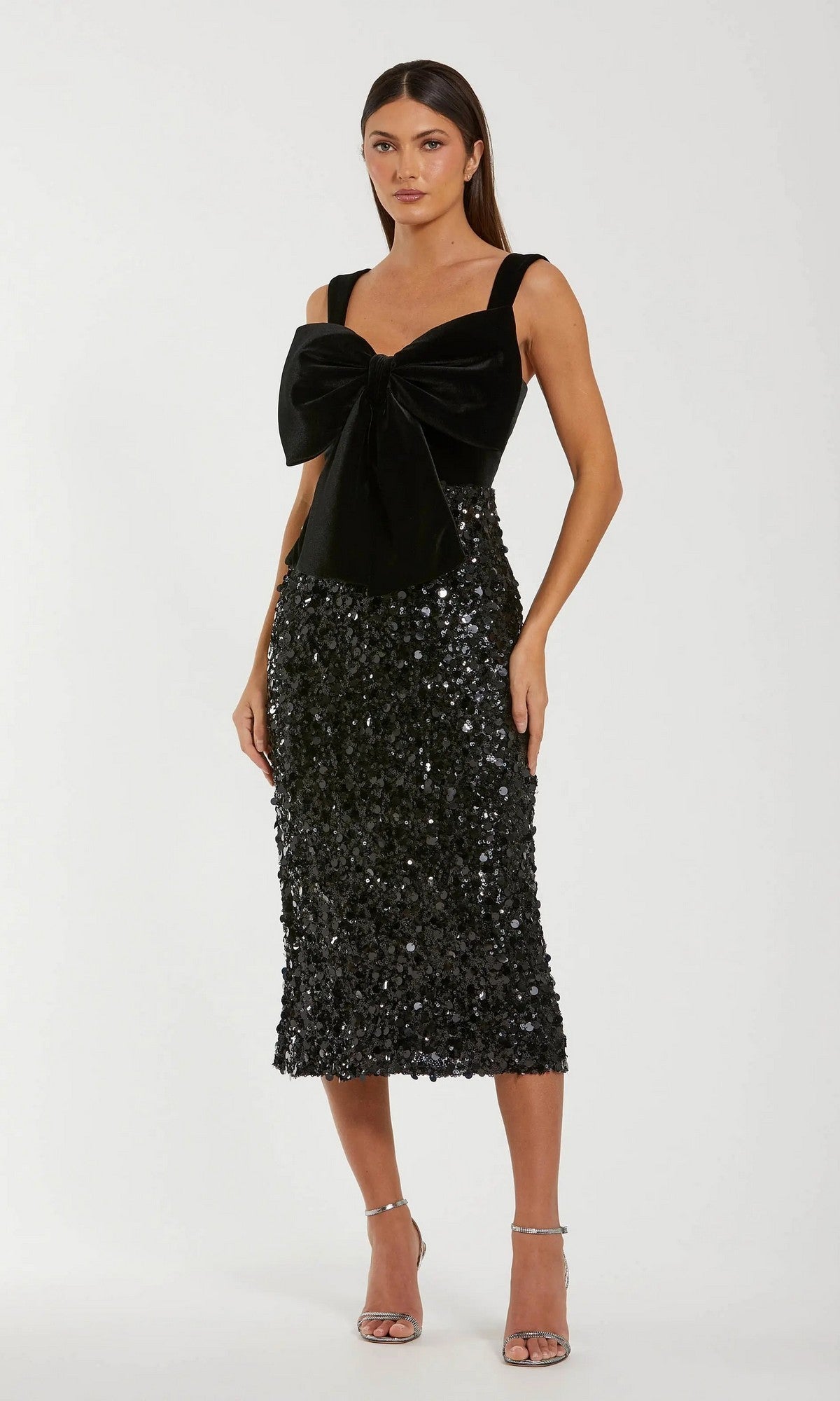 A woman stands in heels against a plain background, wearing the Mac Duggal 27777 midi semi-formal dress featuring a black sleeveless bodice with a large velvet bow and a sparkling sequin skirt.